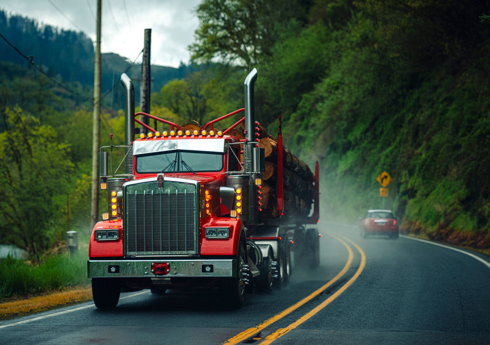 Red semi-truck carrying logs on a misty Oregon highway surrounded by lush greenery.
